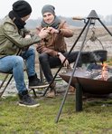 Twee mannen met winterkleding, mutsen en sjaals, toasten met bierflessen bij een kampvuur.