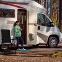 Een vrouw met zonnebril leunt tegen een camper, met een zwarte campingstoel ernaast.