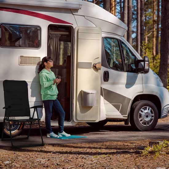 Een vrouw met zonnebril leunt tegen een camper, met een zwarte campingstoel ernaast.