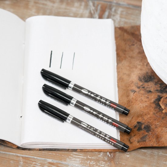 Drie zwarte kalligrafiestiften op een open notitieboek met lijnen getrokken.