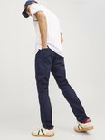 Man met donkere jeans en witte polo, zittend met hand in zak.