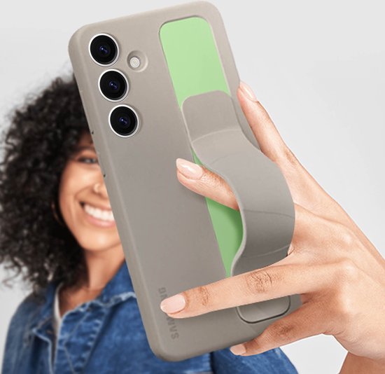 Hand die een beige telefoonhoes met gripband vasthoudt, met een lachende vrouw op de achtergrond.
