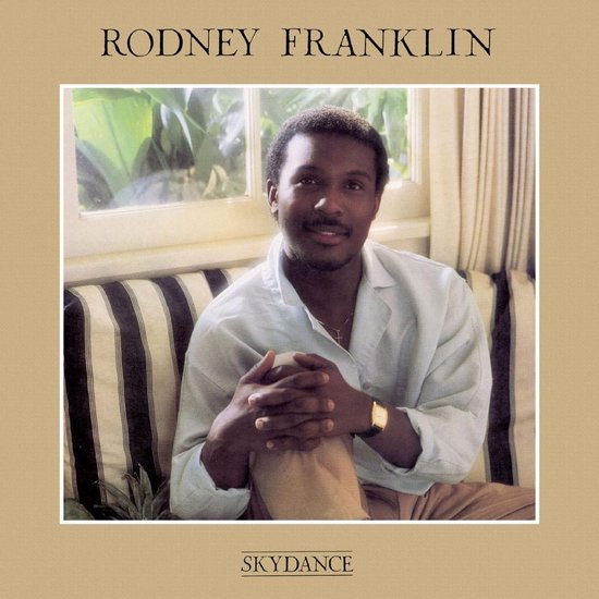 Rodney Franklin zit op een gestreepte bank met gekruiste handen en een lichte blouse.
