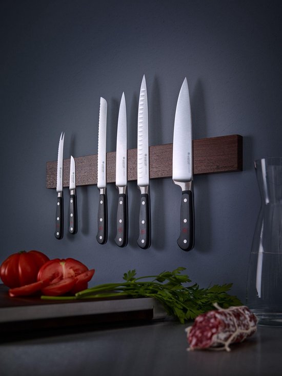 Een set van zes messen met zwarte handvatten hangend aan een magnetische houten strip aan een donkergrijze muur.