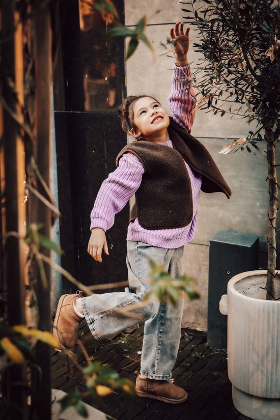 Meisje met opgestoken haar draagt een bruin teddy fleece gilet, paarse trui en spijkerbroek.