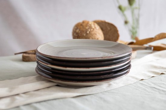 Stapel van vijf grijze dinerborden op een tafel met wit tafelkleed, brood en bladeren op de achtergrond.