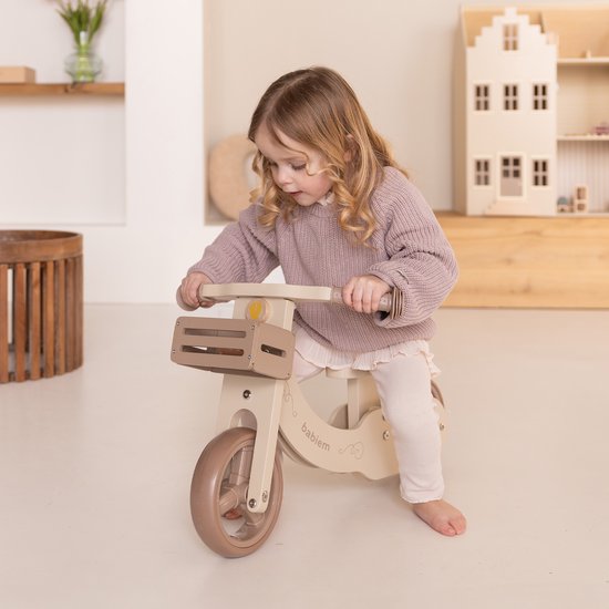 Meisje in gebreide trui op een houten loopfiets, crèmekleurig met bruine details.