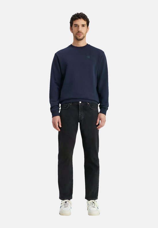 Man met donkerblauwe sweater en zwarte jeans. De sweater heeft een ronde hals en een vierkant logo op de borst.