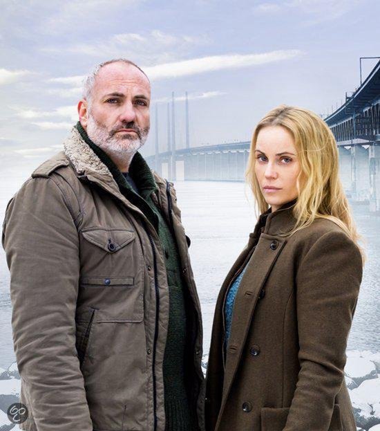 Man met grijze baard in een bruine jas en vrouw met blond haar in een bruine jas, met brug op achtergrond.