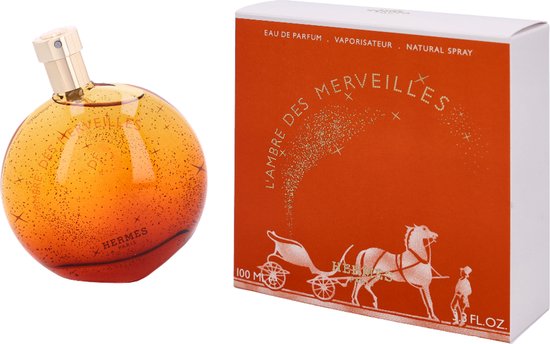Oranje parfumfles met gouden dop, met sprankelende gouden deeltjes en L'Ambre des Merveilles tekst. Naast een oranje doosje met karos en paard.