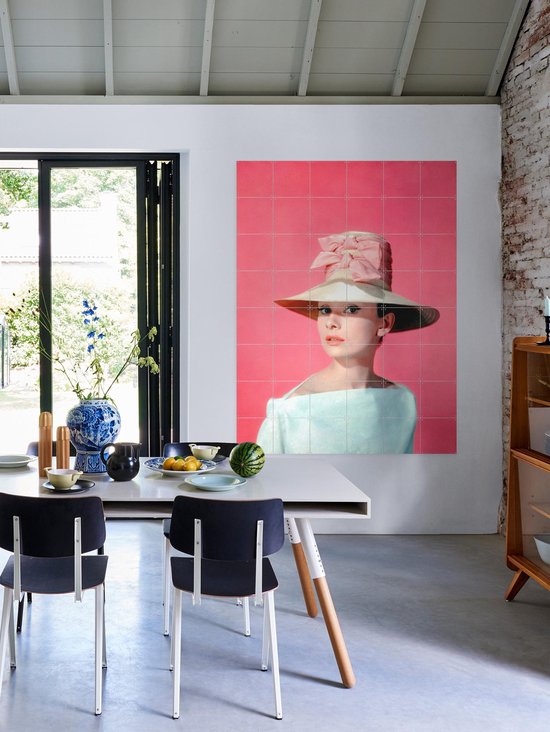 Audrey Hepburn portant un grand chapeau de paille avec un nœud rose, une robe bleu pâle épaules dénudées, sur fond rose, au-dessus d'une table à manger.