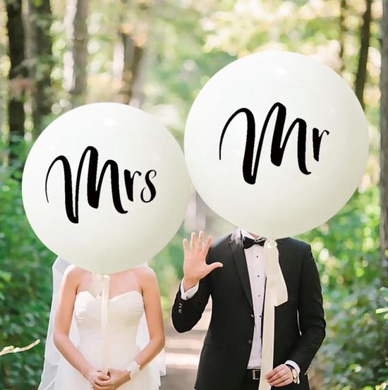 Bruid en bruidegom houden grote witte ballonnen met 'Mrs' en 'Mr' voor hun gezicht in een bosrijke omgeving.