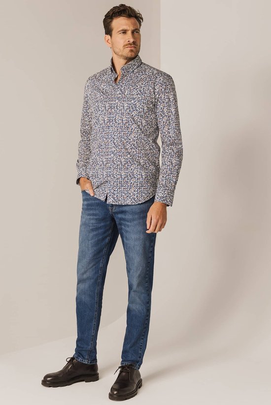 Man met een blauw-wit met bruine print overhemd en spijkerbroek, staand met hand in zak.