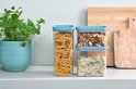 Trois boîtes de conservation transparentes avec couvercles turquoise; une remplie de penne, une de mélange de riz, une de noix.