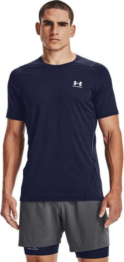 Man in donkerblauw sportshirt met korte mouwen en grijze shorts, staat met grijze schoenen.