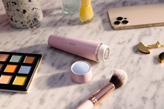 Roze Philips gezichtshaarverwijderaar met LED-ring, naast een make-up palet, kwast, spiegel en telefoon op marmer.