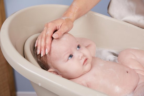 Een hand streelt het hoofd van een baby met blauwe ogen in een beige badje.