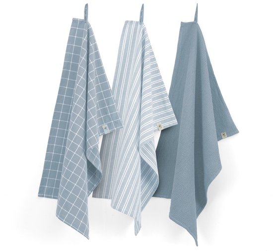 Drie blauwe theedoeken met blok-, streep- en wafelpatroon, hangend aan een witte muur.