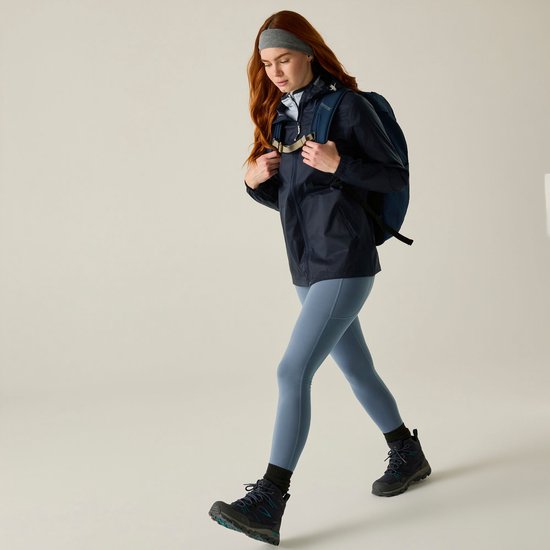 Model met rood haar loopt met een donkerblauwe regenjas, legging en rugzak.