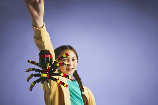 Een meisje met een vlecht houdt een LEGO spin aan een geel koordje boven haar hoofd.