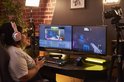Een vrouw met een koptelefoon, zittend aan een bureau met twee monitoren, een toetsenbord en een zwarte audio-controller, klaar om te streamen.