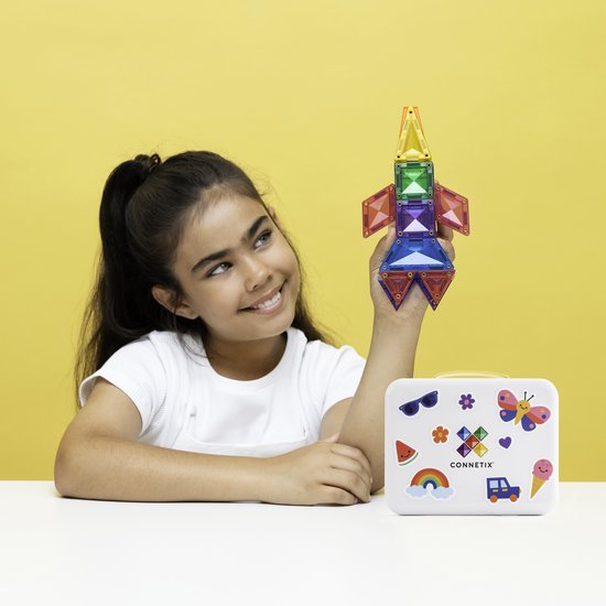 Meisje met een raket gemaakt van Connetix magnetische tegels in regenboogkleuren, naast een witte koffer met stickers.