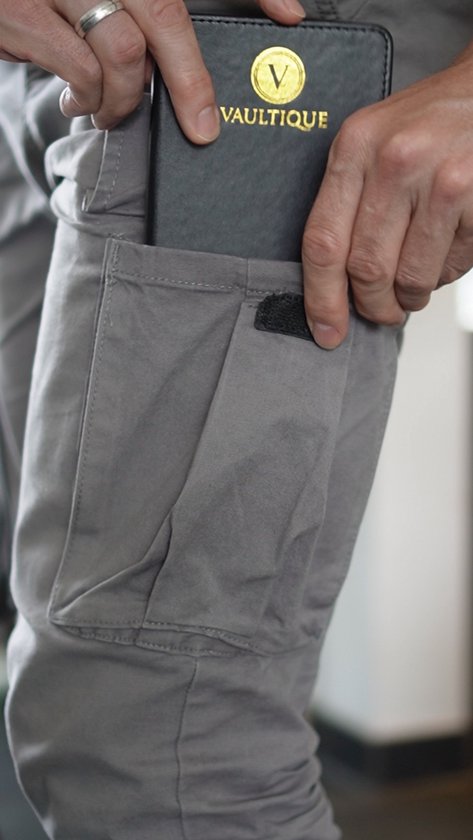 Een hand stopt een zwart wachtwoordenboekje met gouden logo van Vaultique in een zak van een grijze broek.