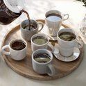 Une verseuse en verre verse du café chaud dans cinq tasses blanches et grises sur un plateau en bois.