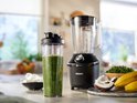 Zwarte Philips blender met een glazen kan, naast een gevuld groen sapje in een meeneembeker, ijsblokjes, bananen en kokos.