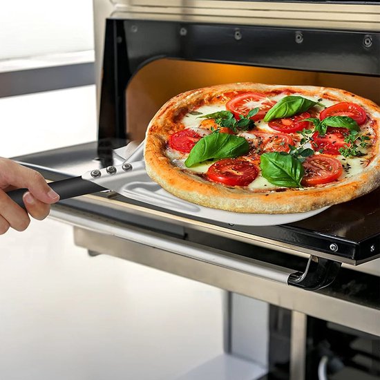 Een hand gebruikt een pizzaschep om een pizza met tomaten en basilicum uit een oven te halen.
