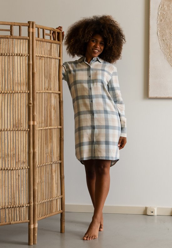 Model met krullend haar draagt een bruin en blauw geruit flanellen nachthemd, staand bij een bamboe kamerscherm.