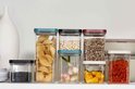 Divers conteneurs alimentaires transparents avec divers remplissages, incluant pâtes, haricots et poivrons.