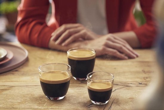 Drie glazen espresso met crèmalaag op een houten tafel, met op de achtergrond een persoon met rode blazer.