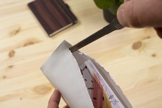 Hand opent een envelop met een briefopener. Een notitieboekje en plant staan ernaast.