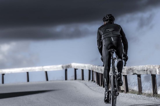 Man fietst op een racefiets op een weg met houten reling, draagt een zwarte jas en fietsbroek.