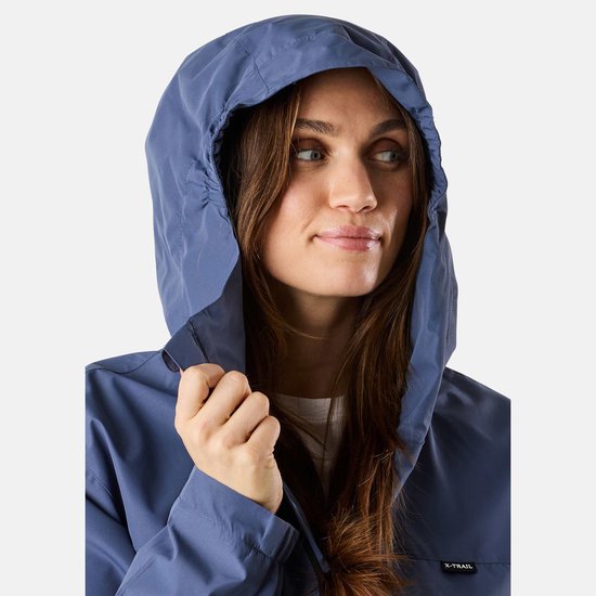 Vrouw met de capuchon van een blauw X-Trail jack op, trekt aan de rand van de capuchon. Zwart X-TRAIL logo op de borst.
