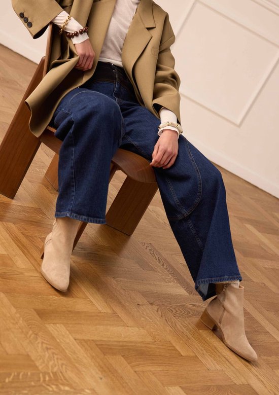 Une femme assise sur une chaise porte une bottine beige en daim à talon bloc.