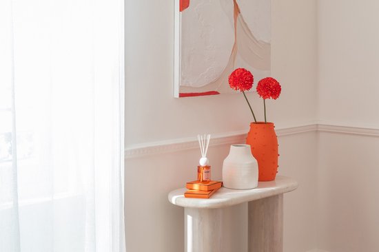 Twee oranje vazen met stippen, één met rode bloemen, naast een witte vaas en diffuser.