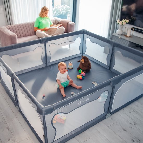 Grijze box met een baby erin, omringd door speelgoed, met een vrouw op de achtergrond.
