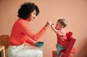 Vrouw met krullend haar in een oranje trui, die een baby in een rode stoel voedt.