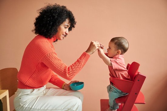 Vrouw met krullend haar in een oranje trui, die een baby in een rode stoel voedt.