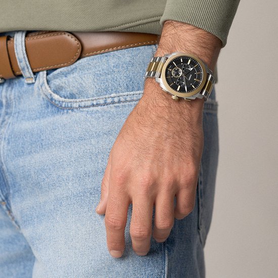 Gouden en zilverkleurig Fossil horloge om een mannelijke pols, gedragen met spijkerbroek en bruine riem.