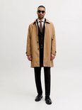 Man in camelkleurige trenchcoat over zwart pak, zonnebril, gestreepte das.