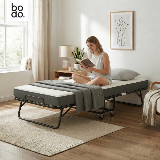 Vouwbed van Bodo met donkergrijs matras, uitgeklapt met een vrouw die leest. Het bed heeft een zwart metalen frame en wieltjes.