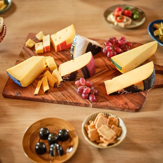 Houten plank met verschillende stukken gele kaas, rode druiven, olijven en crackers.