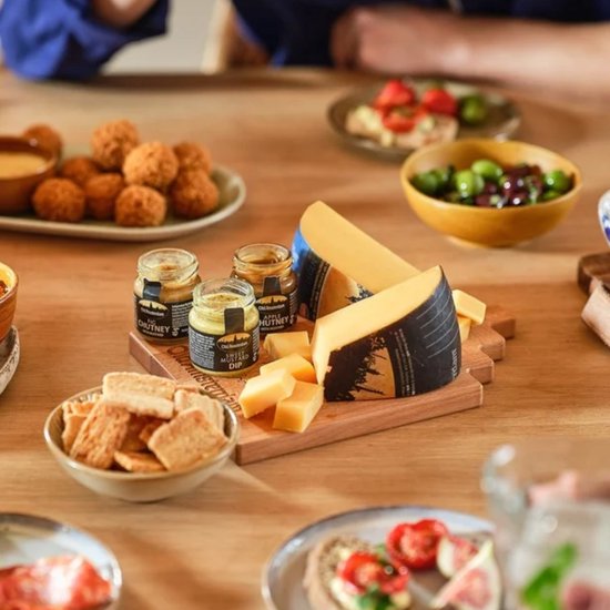 Een houten plank met Old Amsterdam kaas, mini-chutneys, crackers en olijven op een gedekte tafel.