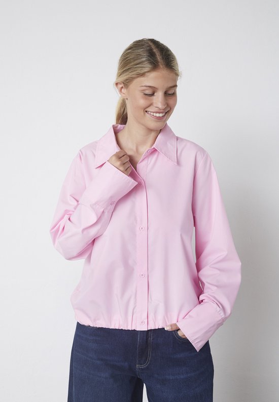 Mannequin aux longs cheveux, portant une blouse rose pâle oversize boutonnée, col rabattu et pantalon.