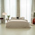 Een bed met een zandkleurig dekbedovertrek en kussens, in een minimalistische ruimte met grote ramen en betonnen pilaren.