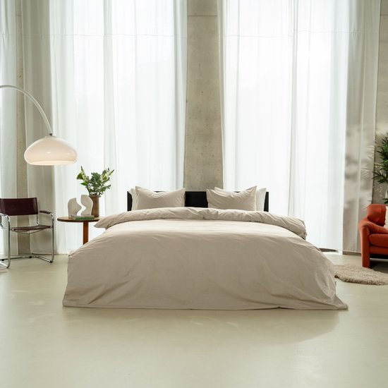 Een bed met een zandkleurig dekbedovertrek en kussens, in een minimalistische ruimte met grote ramen en betonnen pilaren.