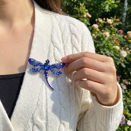 Blauwe libelle broche met zilverkleurig metaal en fonkelende facet geslepen stenen op een witte gebreide cardigan.